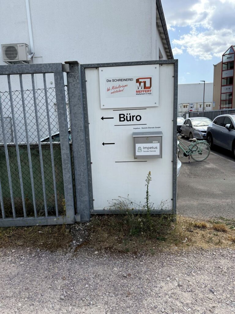 Tor mit Wegweiser „Büro“ und Impetus-Schild, dahinter der Parkplatz