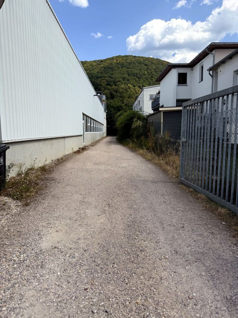 Schotterweg zwischen zwei Hallen im Gewerbegebiet Schriesheim Süd, dahinter der bewaldete Odenwald